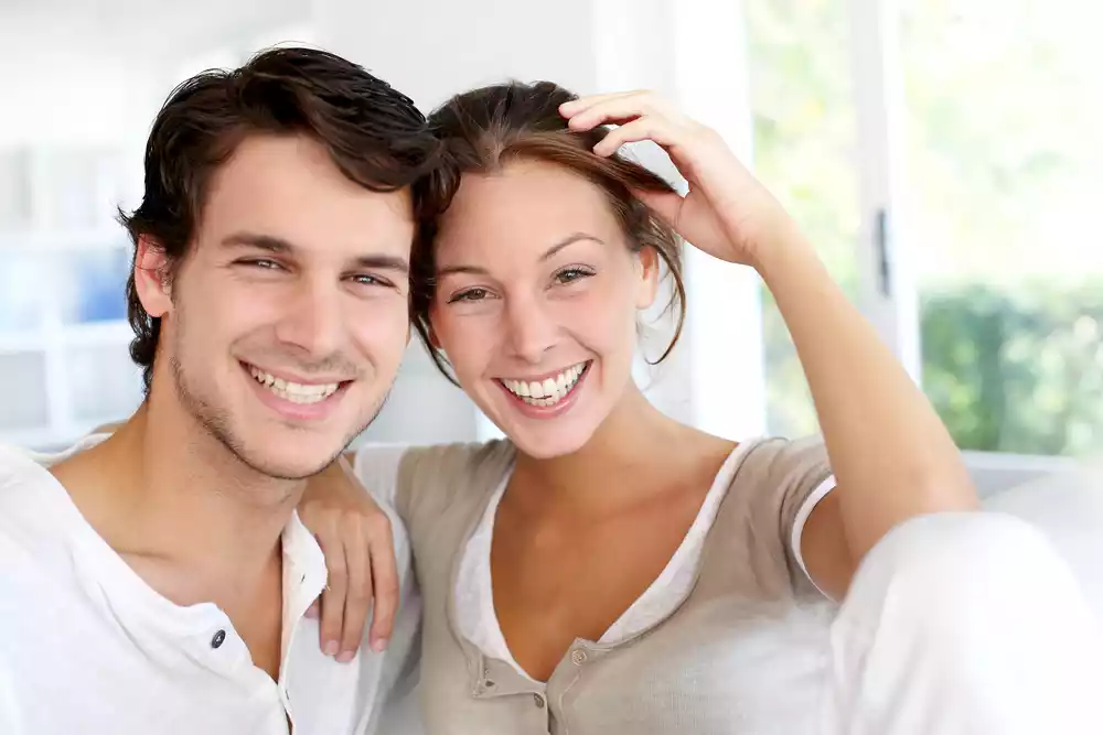 A happy young couple sitting close and smiling at the camera, in a brightly lit room.