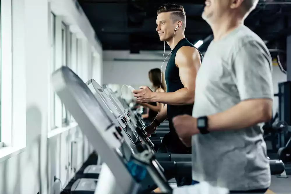 Two men jogging on treadmills in a modern gym, focusing on their workout with a window in the background.