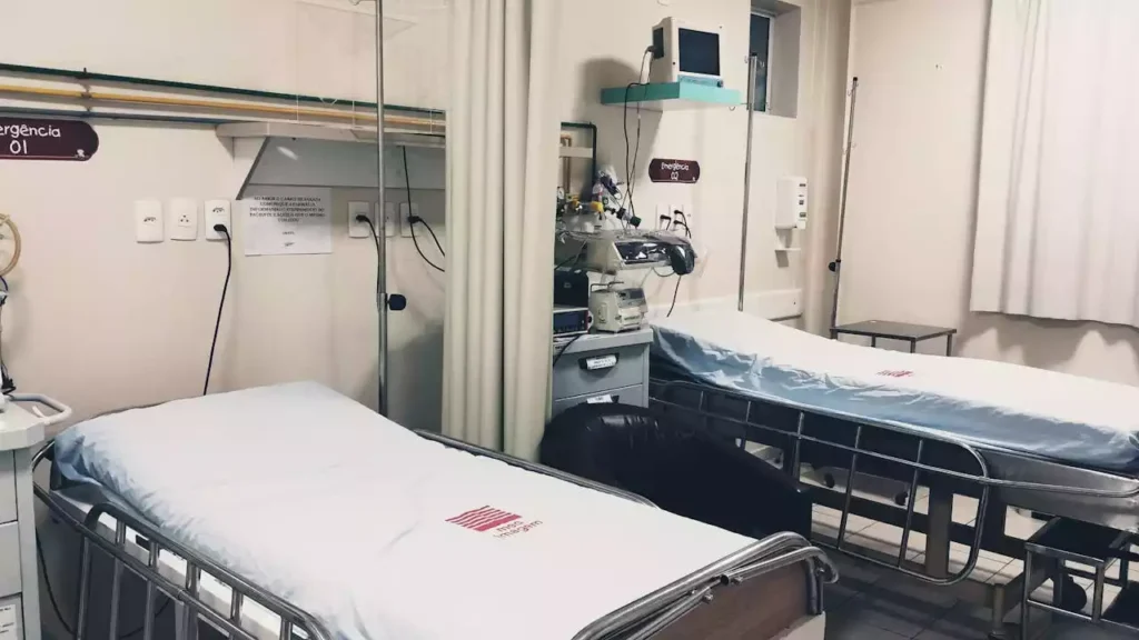 Hospital room with two empty beds, medical equipment, and a curtain divider. Walls have mounted devices and signs.
