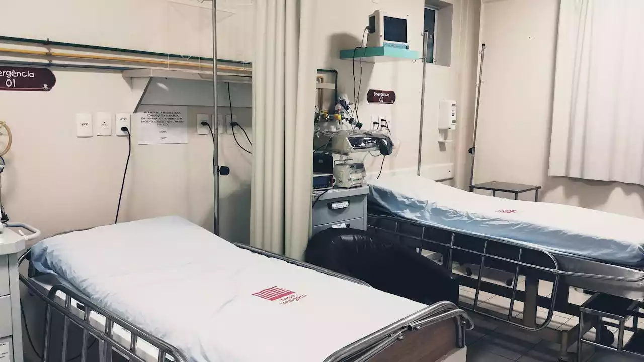 Hospital room with two empty beds, medical equipment, and a curtain divider. Walls have mounted devices and signs.