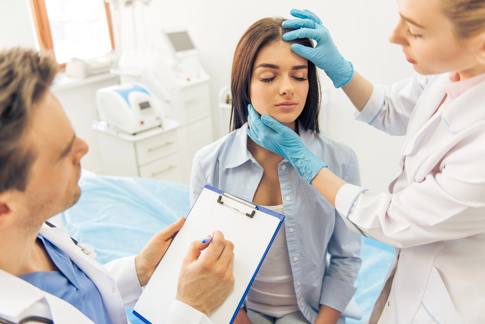 Two doctors examine a young woman’s face in a medical office, discussing potential blepharoplasty benefits. One takes notes on a clipboard while the other, both wearing gloves, gently touches the patient’s chin and forehead.