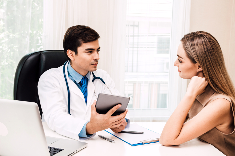 A doctor holding a tablet discusses blepharoplasty benefits with a woman at a desk, where a laptop, clipboard, and pen are arranged in a well-lit office.