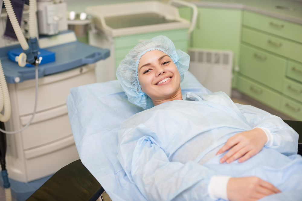 A person in a surgical gown and cap is lying on a hospital bed, smiling at the camera, possibly experiencing blepharoplasty benefits in a medical setting.