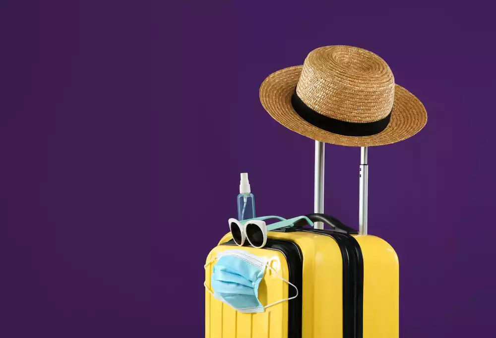 Yellow suitcase with a straw hat, sunglasses, face mask, and sanitizer bottle against a purple background—perfect essentials for turismo médico en México.