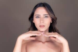 A woman with long brown hair poses against a plain background, holding her hands under her chin and looking directly at the camera.
