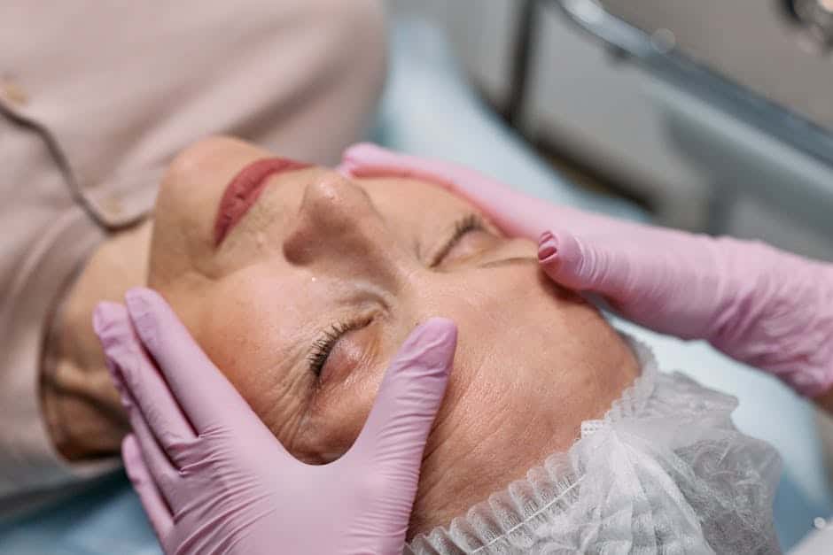 A person wearing a hair cover receives a facial massage from someone wearing pink gloves, lying on a treatment bed.