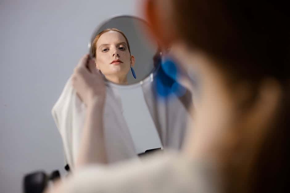 Person holding a round mirror, showing their face reflected in it, with a neutral expression and blue earring visible.