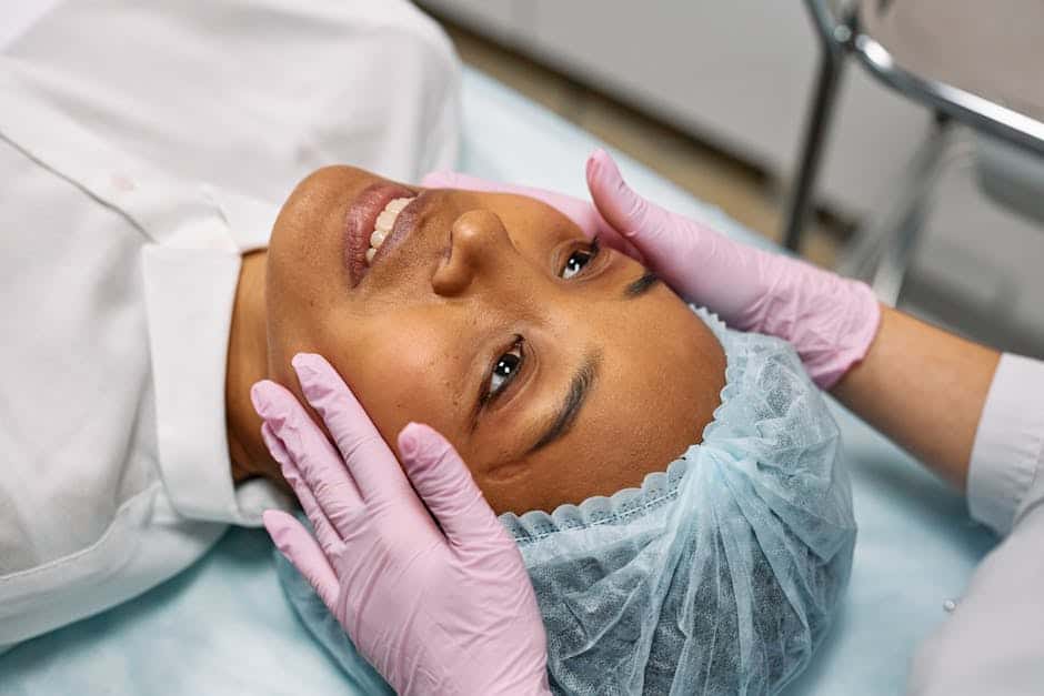 Person lying on a medical bed, wearing a hair cover, while another person in pink gloves examines their face in a clinical setting.