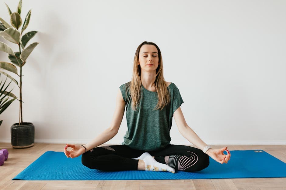 A woman sits cross-legged on a blue yoga mat indoors, meditating with eyes closed and hands resting on her knees.