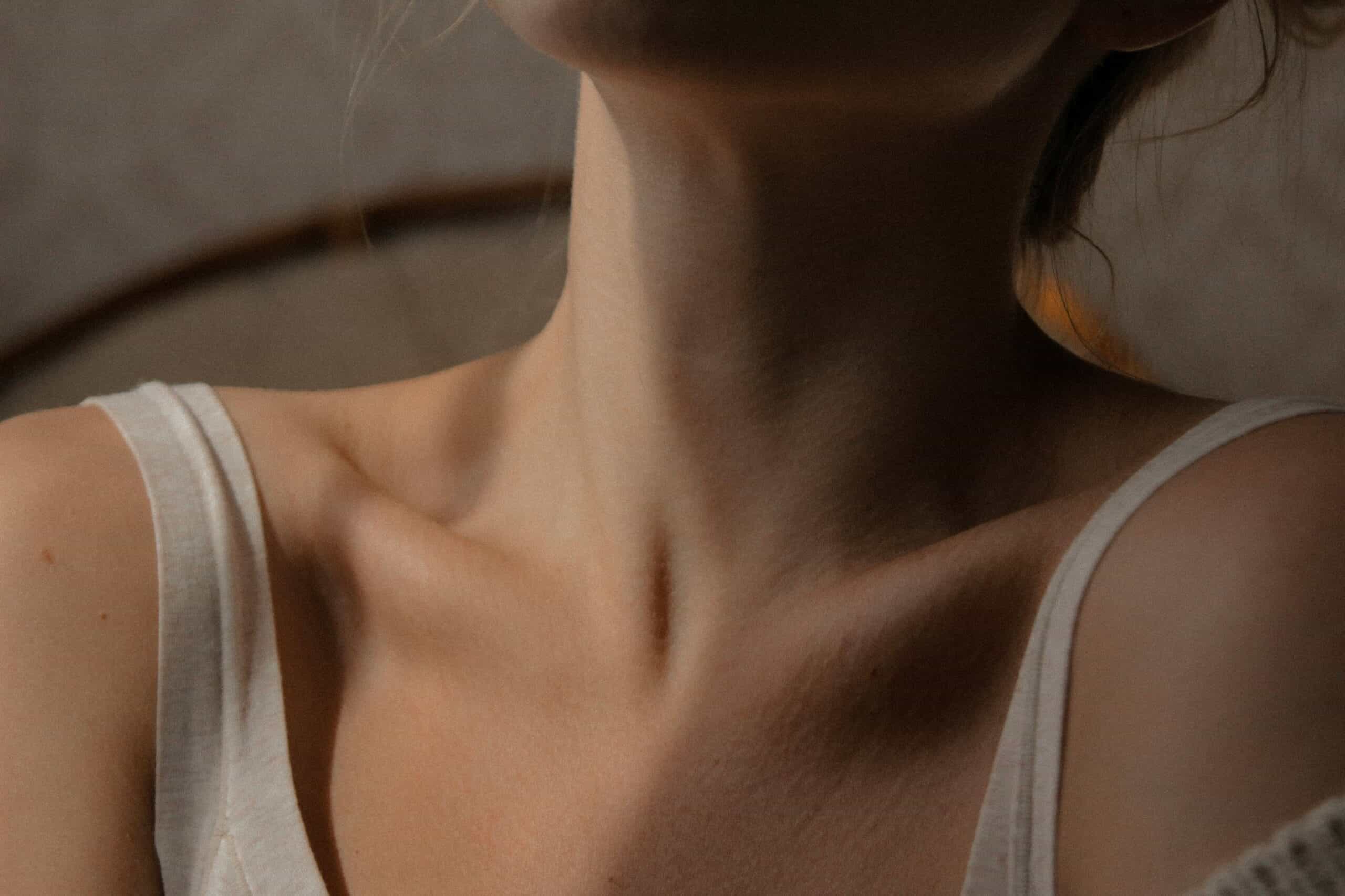 Close-up of a person's neck, collarbone, and upper chest wearing a white tank top, with soft lighting and a neutral background.
