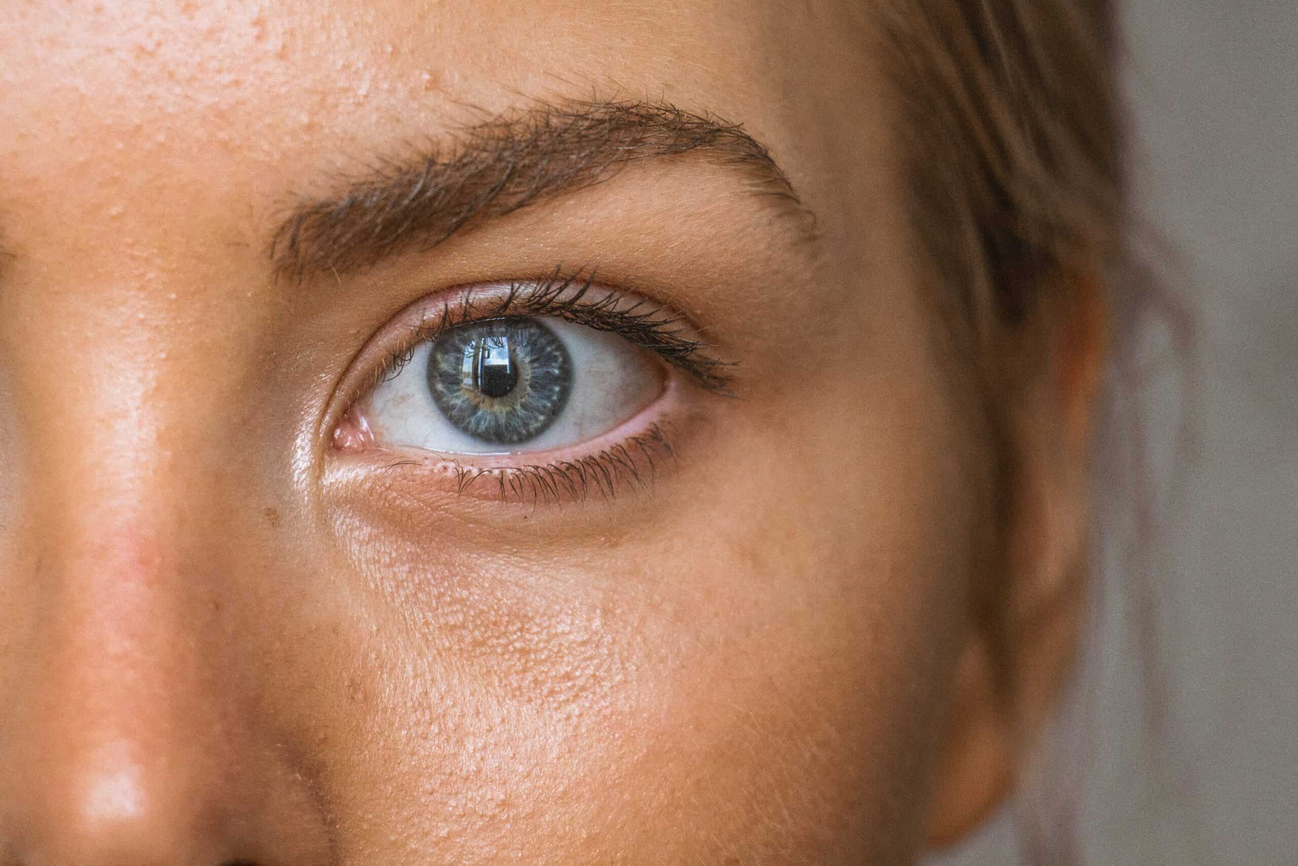 Close-up of a person's blue eye, eyebrow, and part of their nose and cheek, showing detailed skin texture and natural light reflection in the eye.