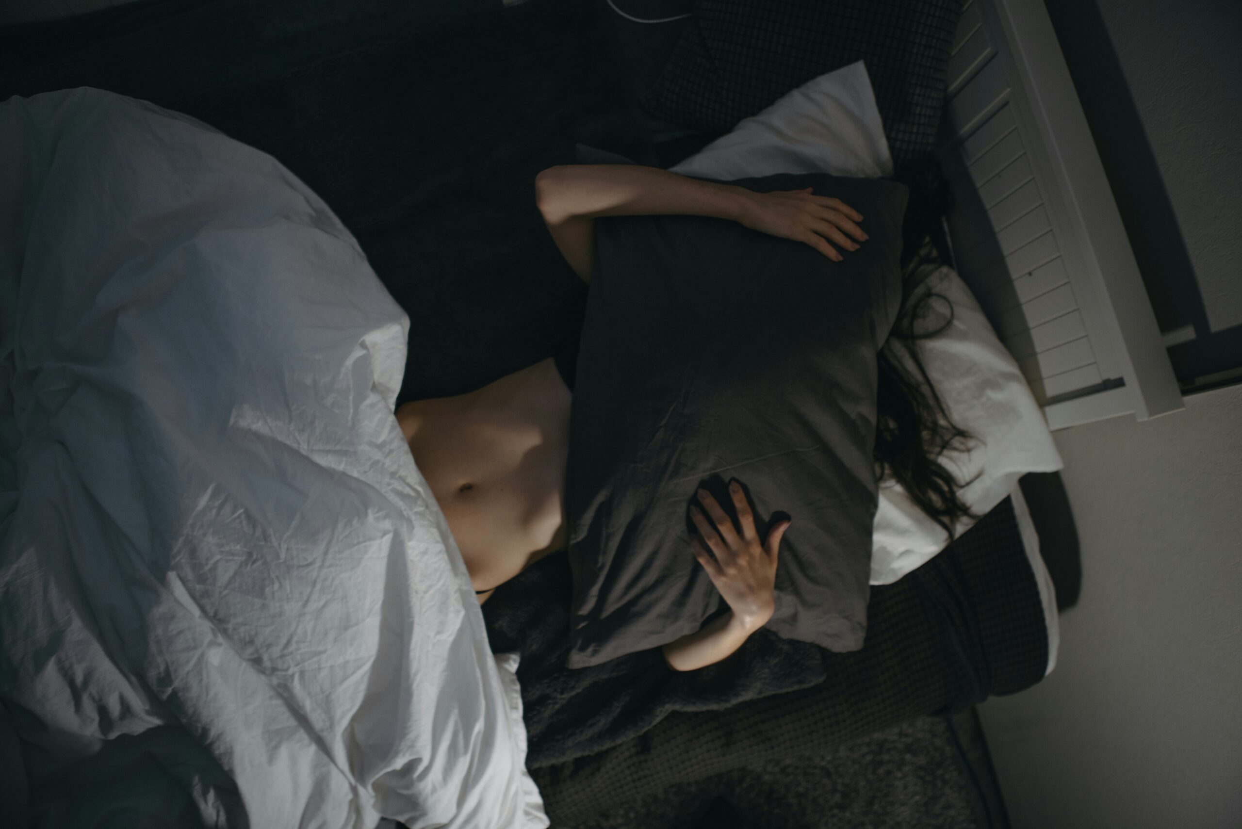 A person lying in bed, partially covered by a white blanket, holds a dark pillow over their face and torso.