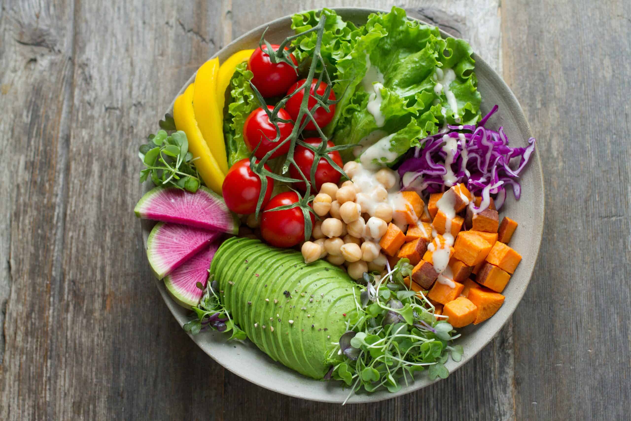 A bowl filled with avocado, chickpeas, cherry tomatoes, lettuce, yellow bell pepper, sweet potato, watermelon radish, microgreens, shredded cabbage, and dressing.