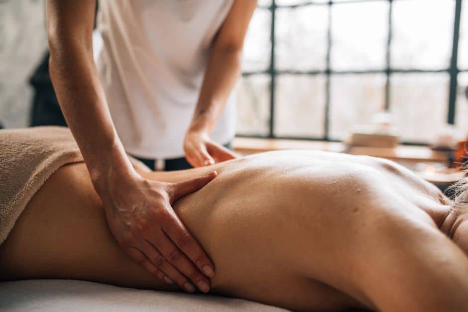 A person receives a back massage from a massage therapist in a sunlit room, with hands applying pressure to the client’s back.