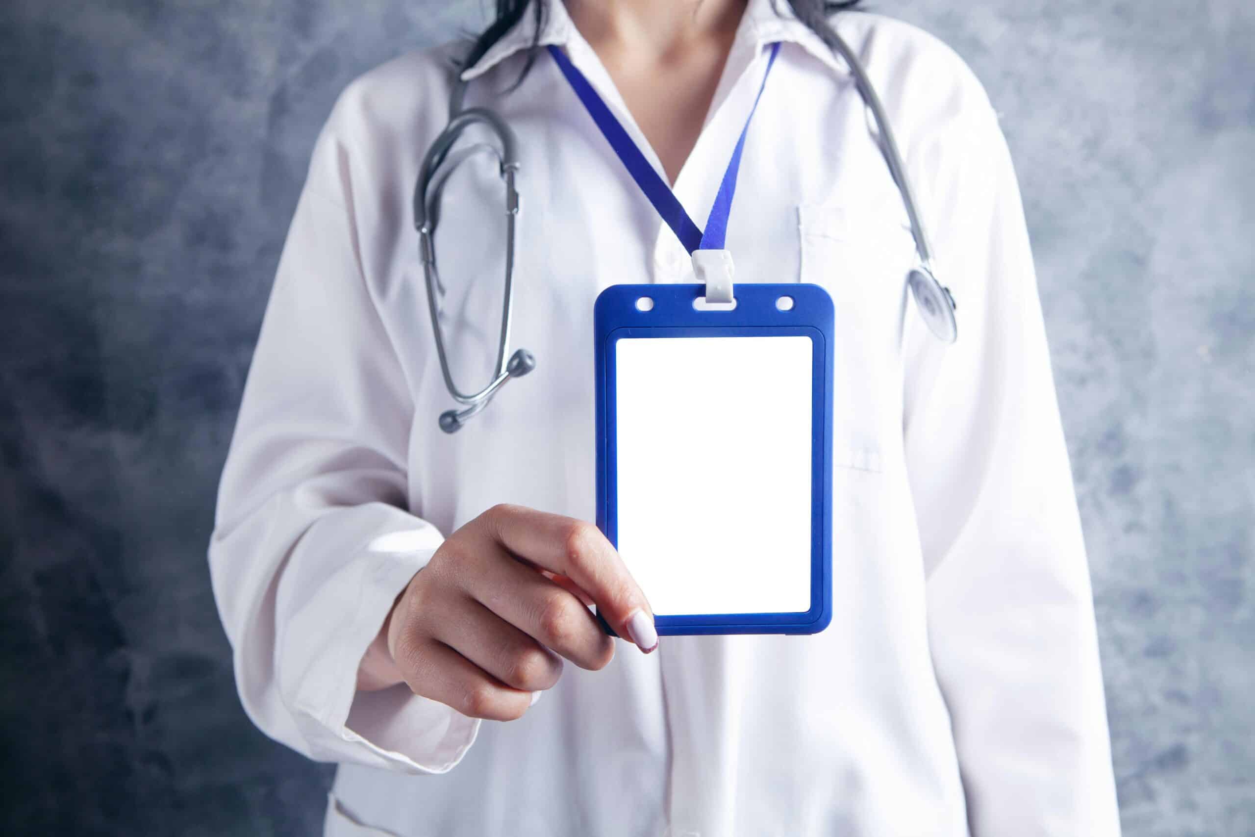 A person in a white lab coat with a stethoscope displays a blank ID badge against a gray background.