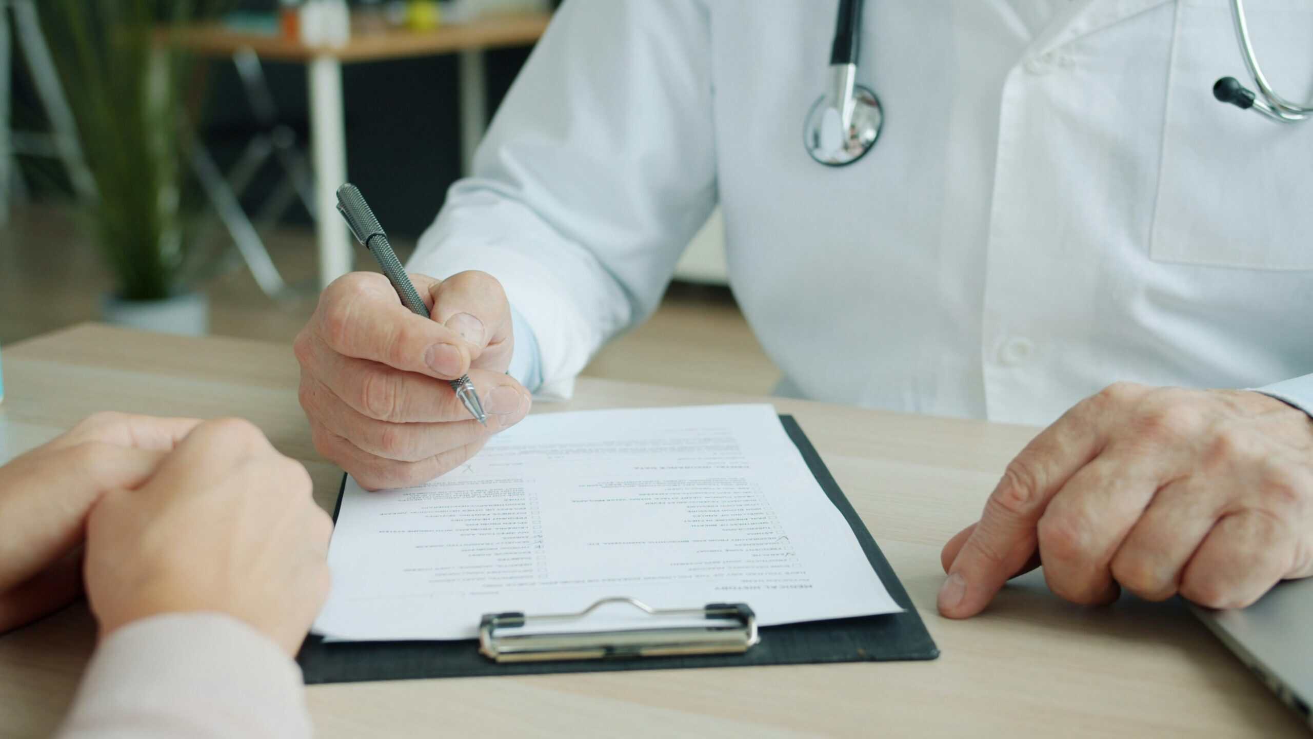 A doctor with a stethoscope discusses a medical form with a patient, holding a pen and pointing to the document on a clipboard.